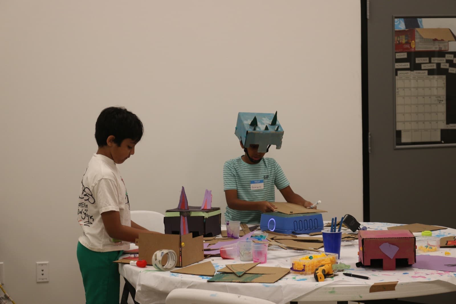 Camper in a handmade cardboard dragon helmet crafting at the build table